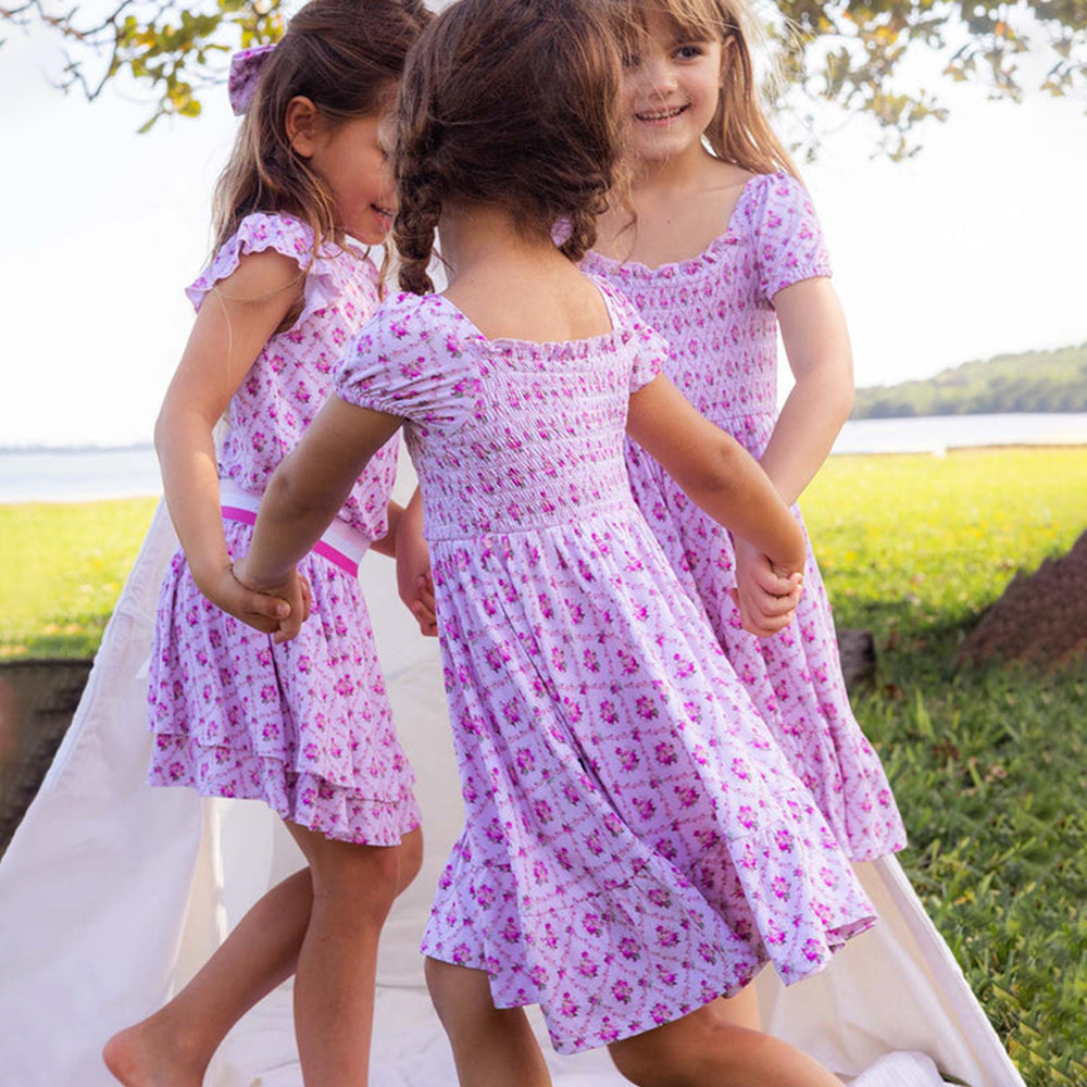 Three young girls in matching pink floral dresses standing outdoors near a body of water.