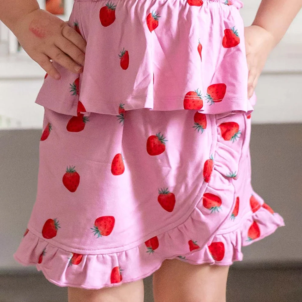 A close-up of a child wearing a pink tiered skirt with a strawberry print, showing the ruffled hem and side pockets.