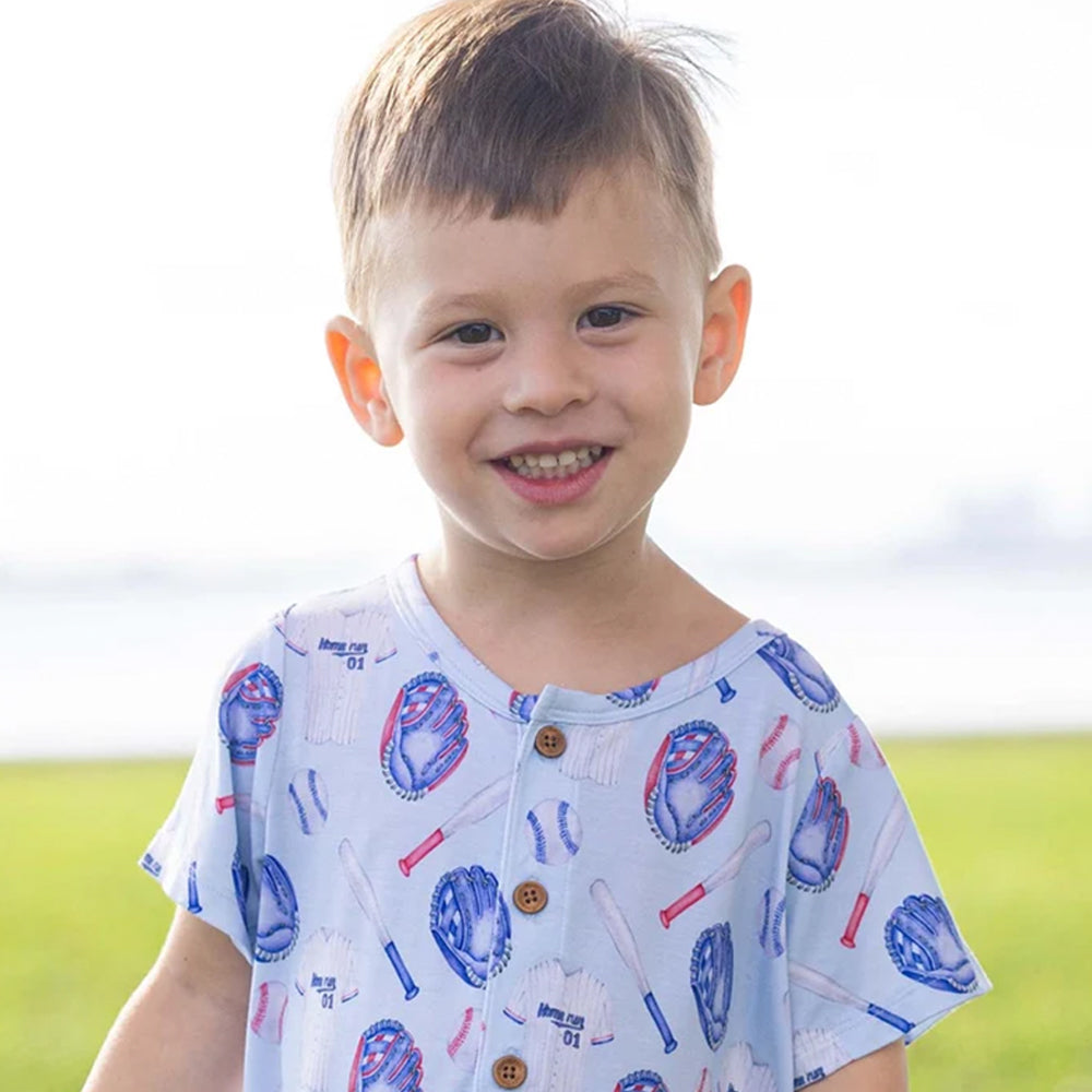 A young boy smiling outdoors, wearing a light blue short-sleeve henley shirt with a baseball-themed print featuring blue and red baseballs, bats, and white uniforms.