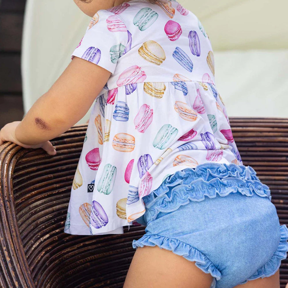Child wearing a colorful macaron-patterned shirt and blue shorts sitting on a wicker chair.
