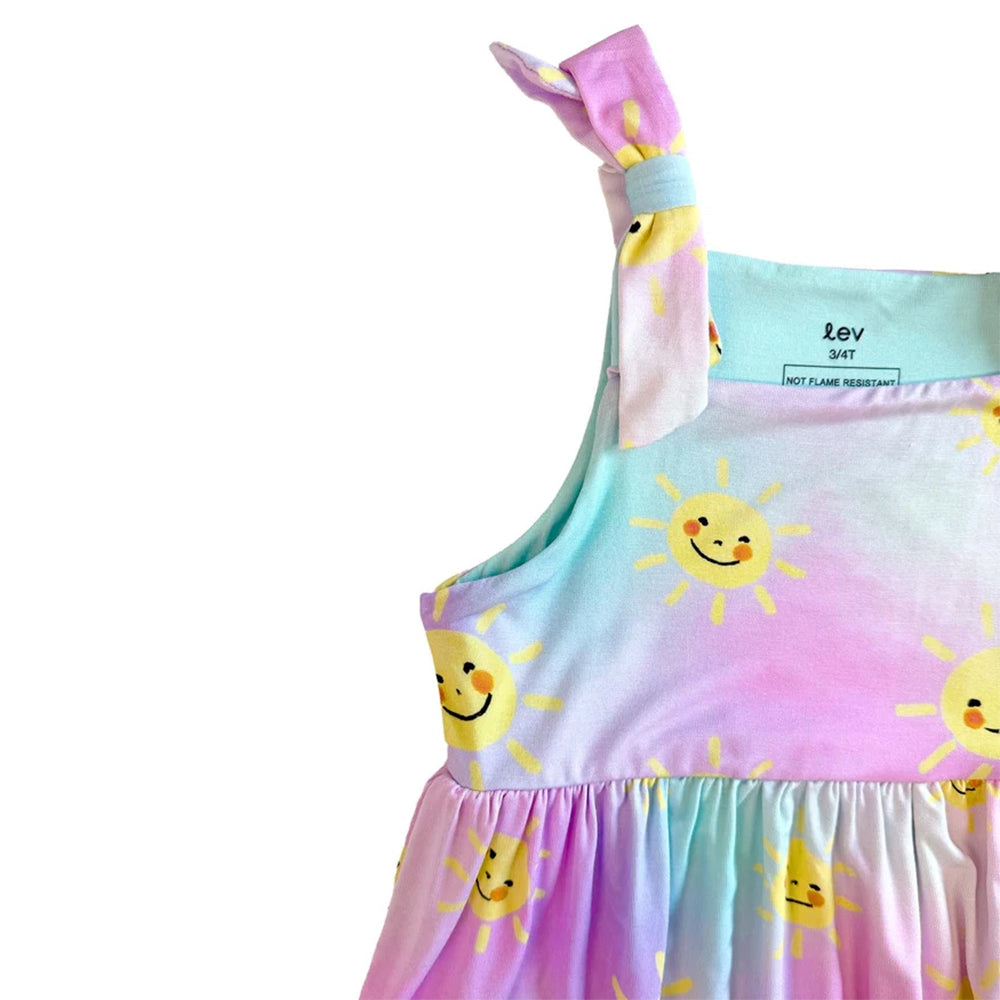 Close-up of a children's dress with a pastel tie-dye gradient in pink, blue, and purple, featuring a yellow smiley sun print and a knotted shoulder strap, shown on a white background.