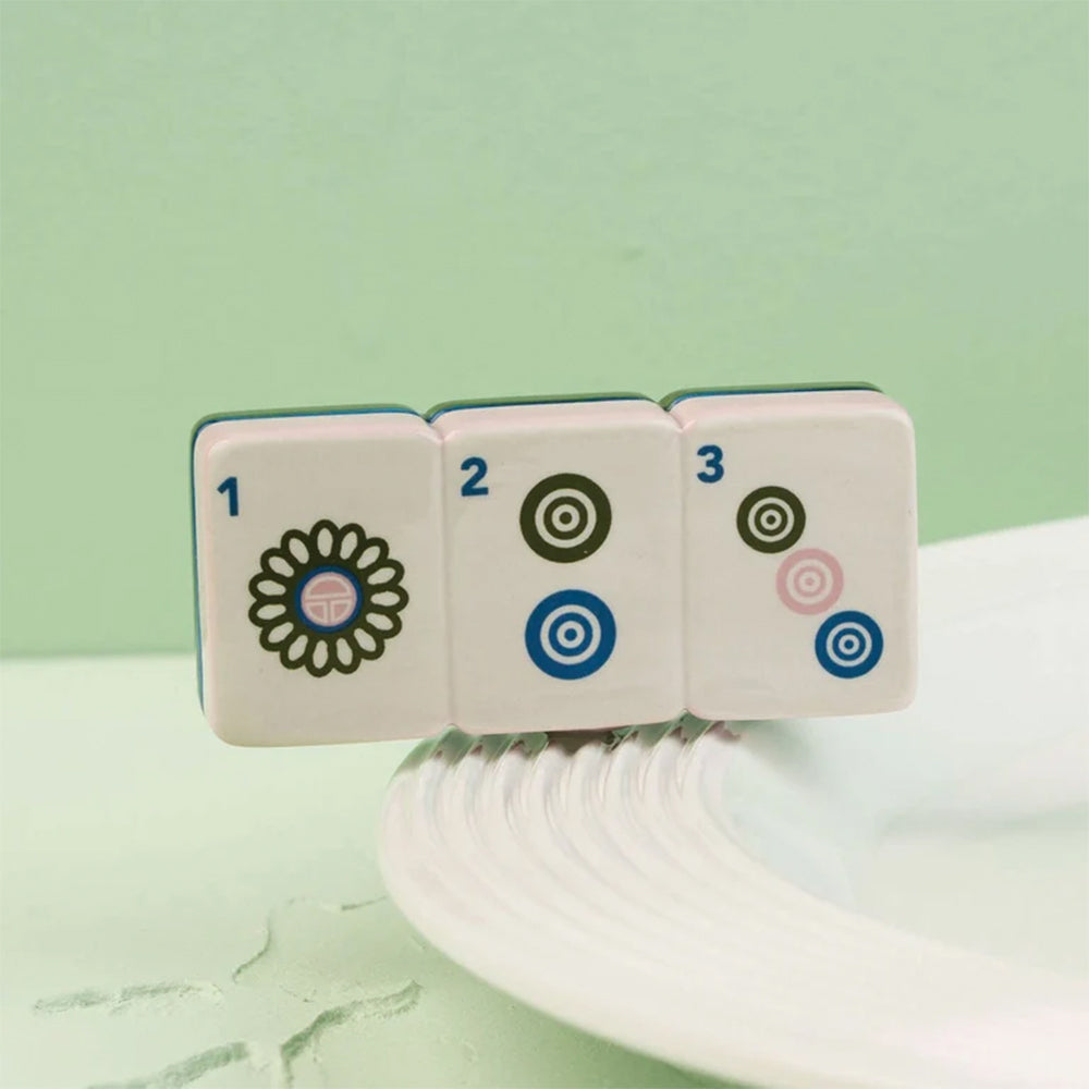 Three white mahjong tiles with black, blue, and pink symbols resting on the edge of a white plate against a pale green background.