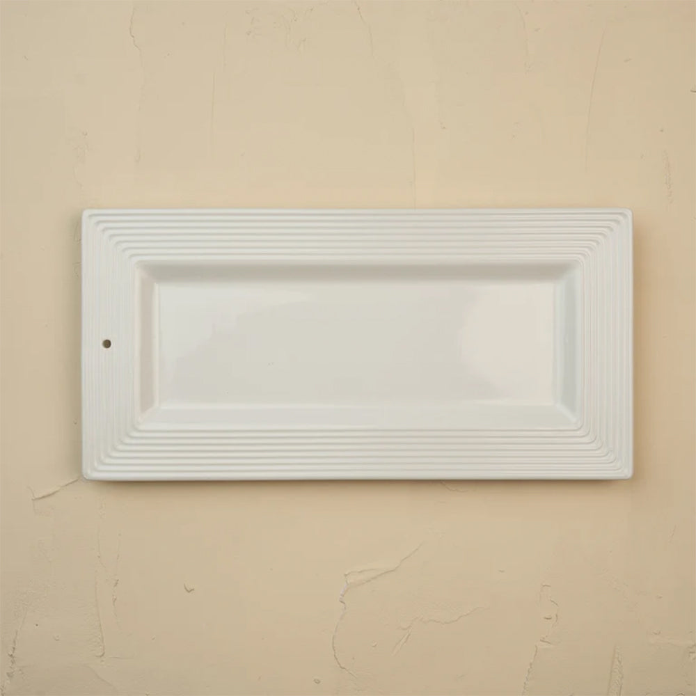 A white rectangular ceramic serving platter with a fluted border, shown against a textured beige wall.