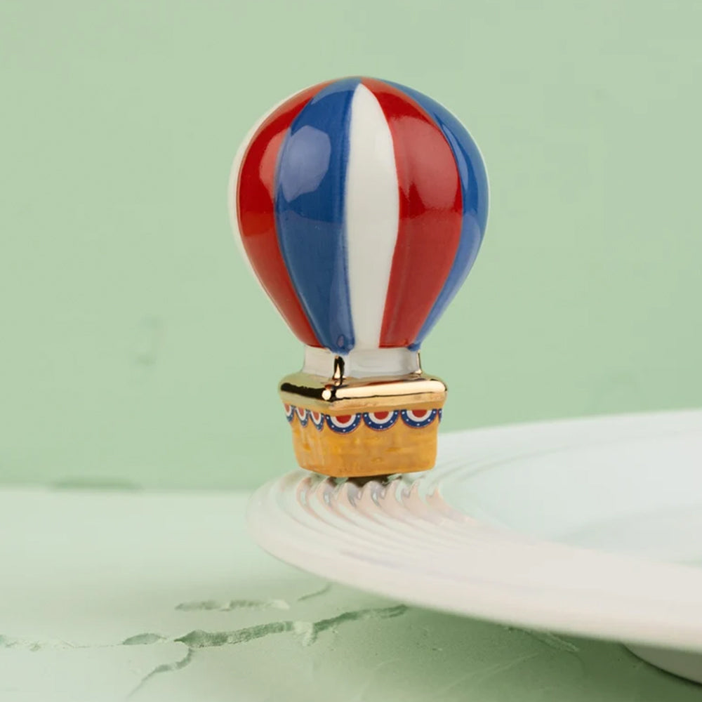 A small decorative hot air balloon charm or bead with red, white, and blue vertical stripes and a gold-toned basket, displayed on a white plate against a pale green background.