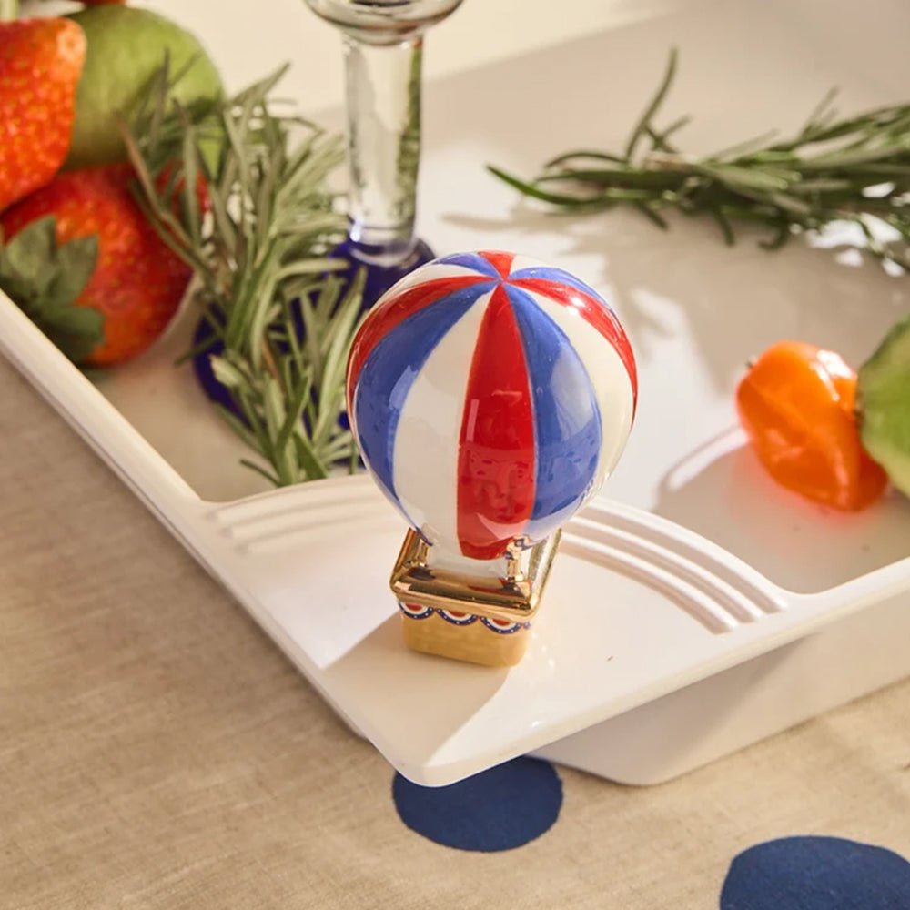 A small ceramic hot air balloon ornament with red, white, and blue stripes and a gold base, displayed on a white tray with strawberries, rosemary, and other decorative items.