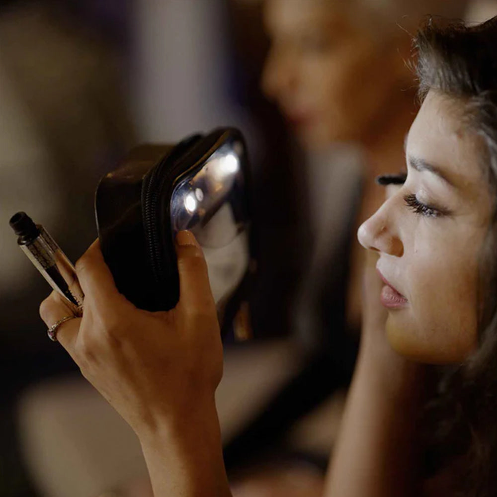 Woman applying makeup with a compact mirror and brush in a blurred indoor setting