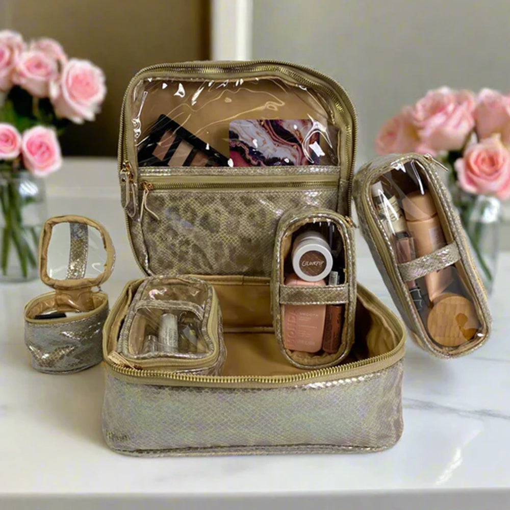 Gold travel makeup case with cosmetic items on a white surface with pink flowers in the background