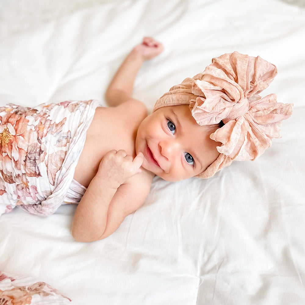 A baby lying on white bedding, wearing a dusty pink fabric headband with a large bow and a matching floral-patterned wrap.