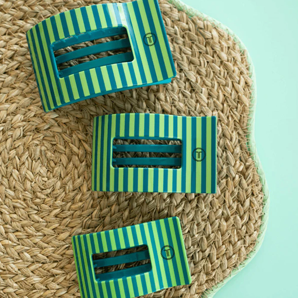 Three green and blue striped hair clips on a woven mat with a light blue background