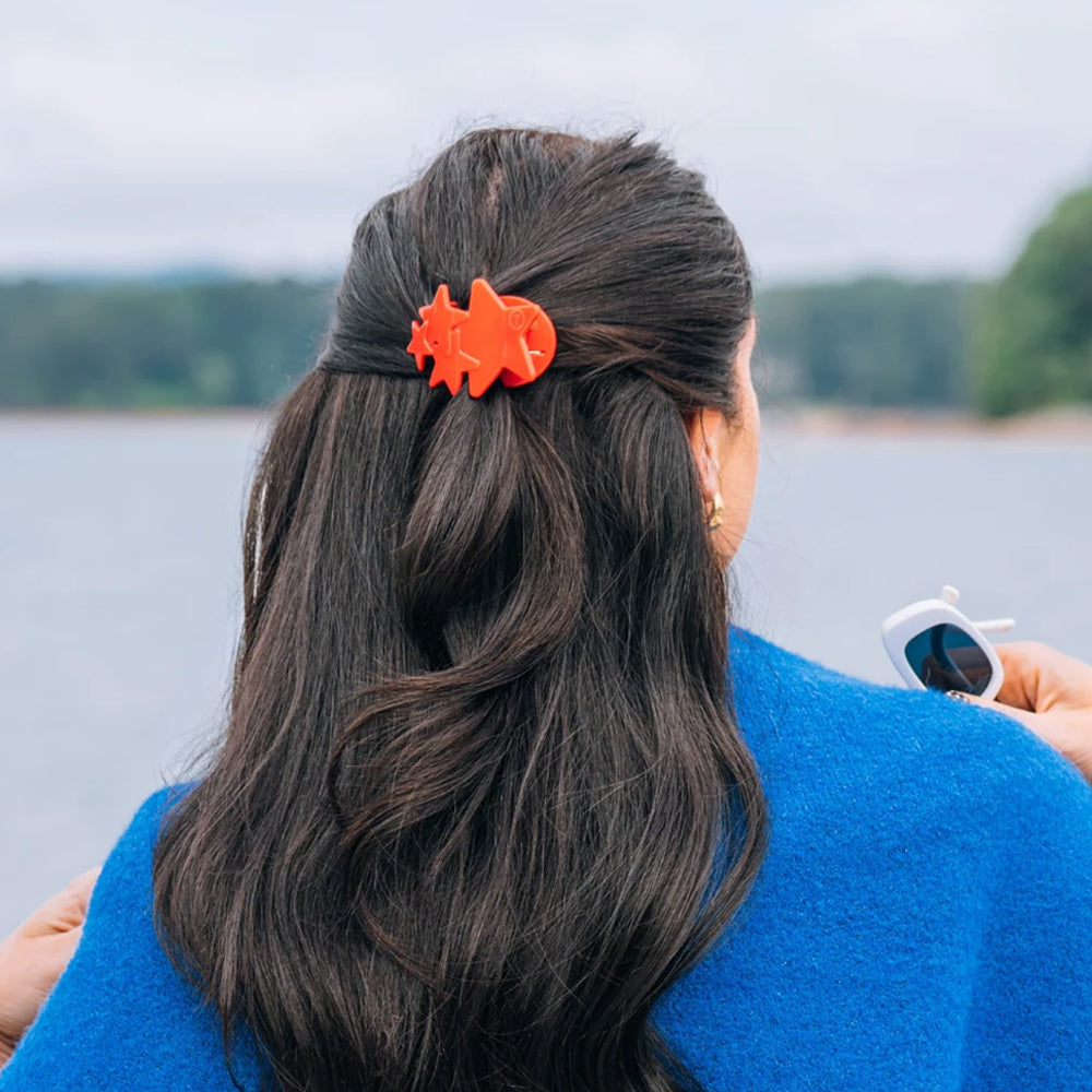 Person with an red hair clip in their dark hair, standing by a body of water.