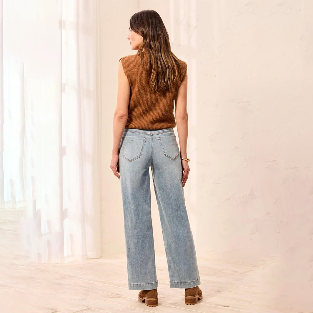 A woman is shown from behind wearing a brown ribbed knit sleeveless sweater and light-wash wide-leg denim jeans with decorative stitching on the back pockets, standing in a bright, neutral indoor setting.