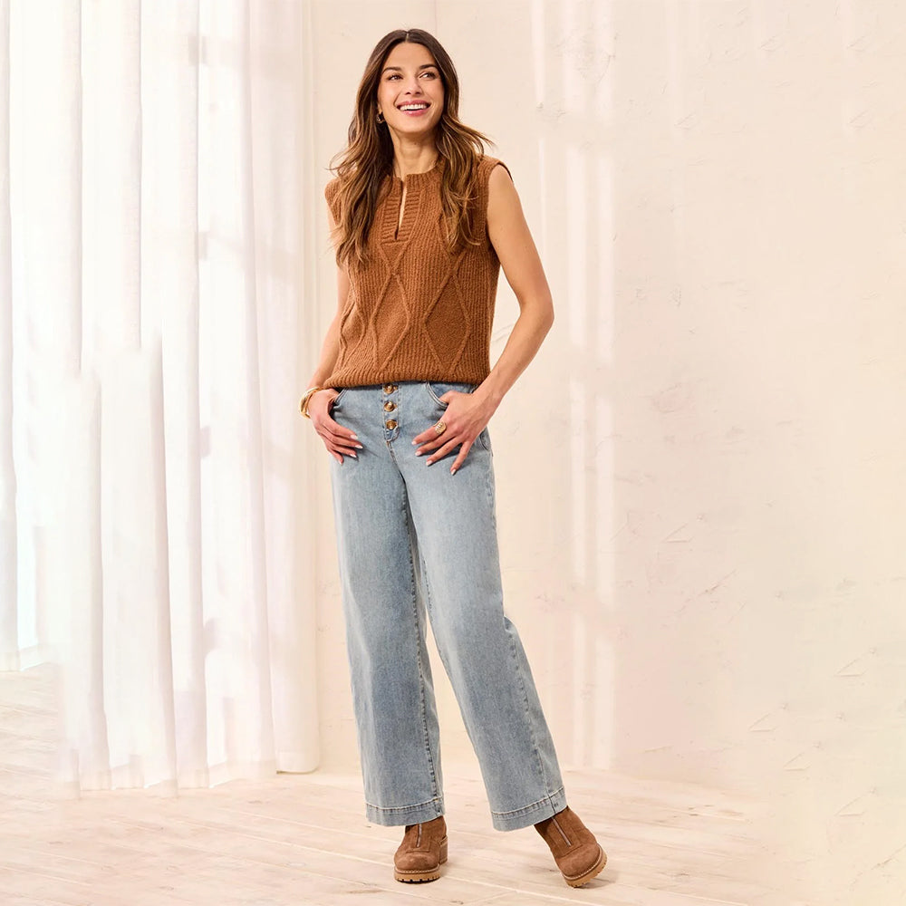 A woman modeling a sleeveless brown cable-knit vest with a zipper neckline, paired with light-wash wide-leg jeans and brown suede boots, standing in a brightly lit room with sheer curtains.