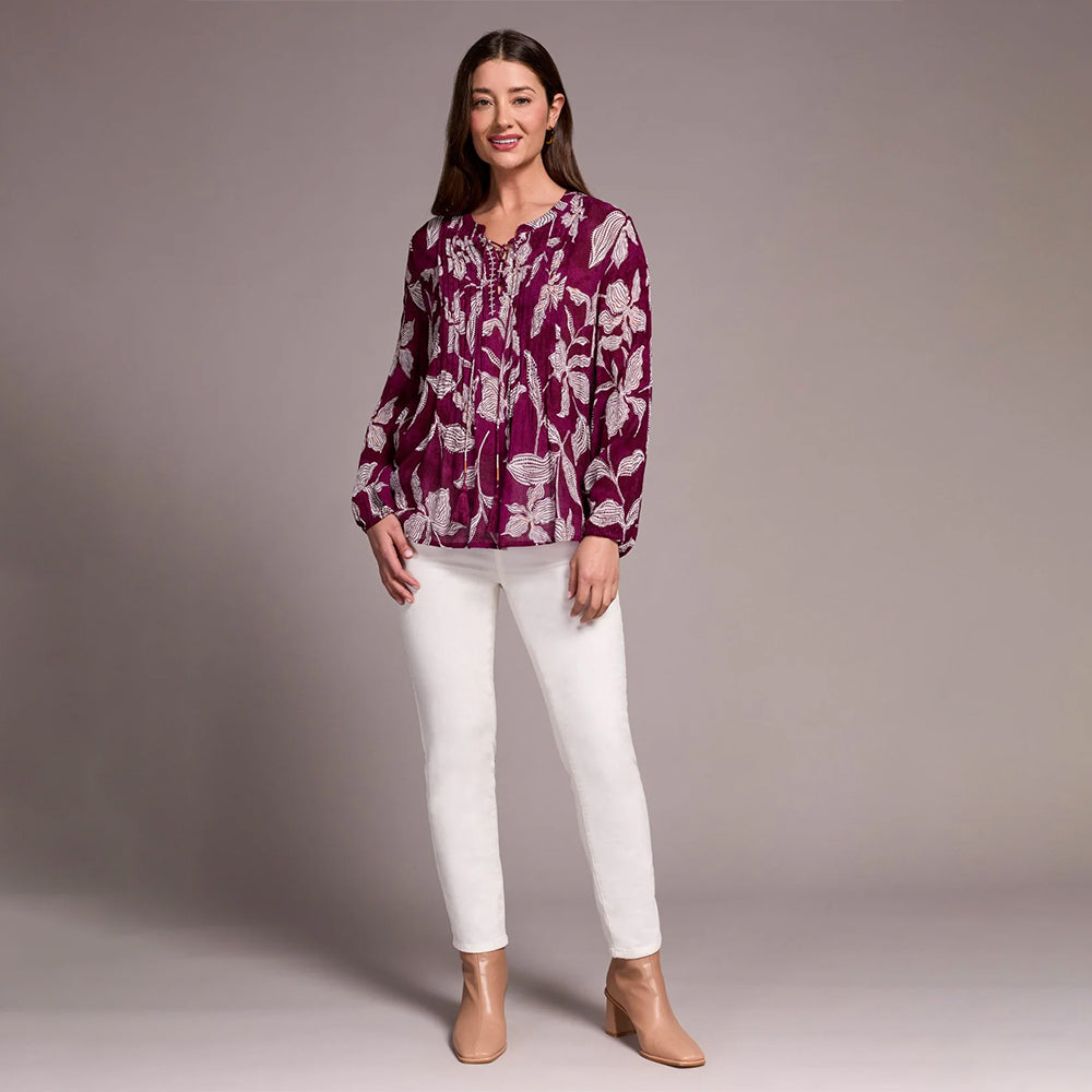 Woman wearing a purple floral blouse and white pants against a gray background