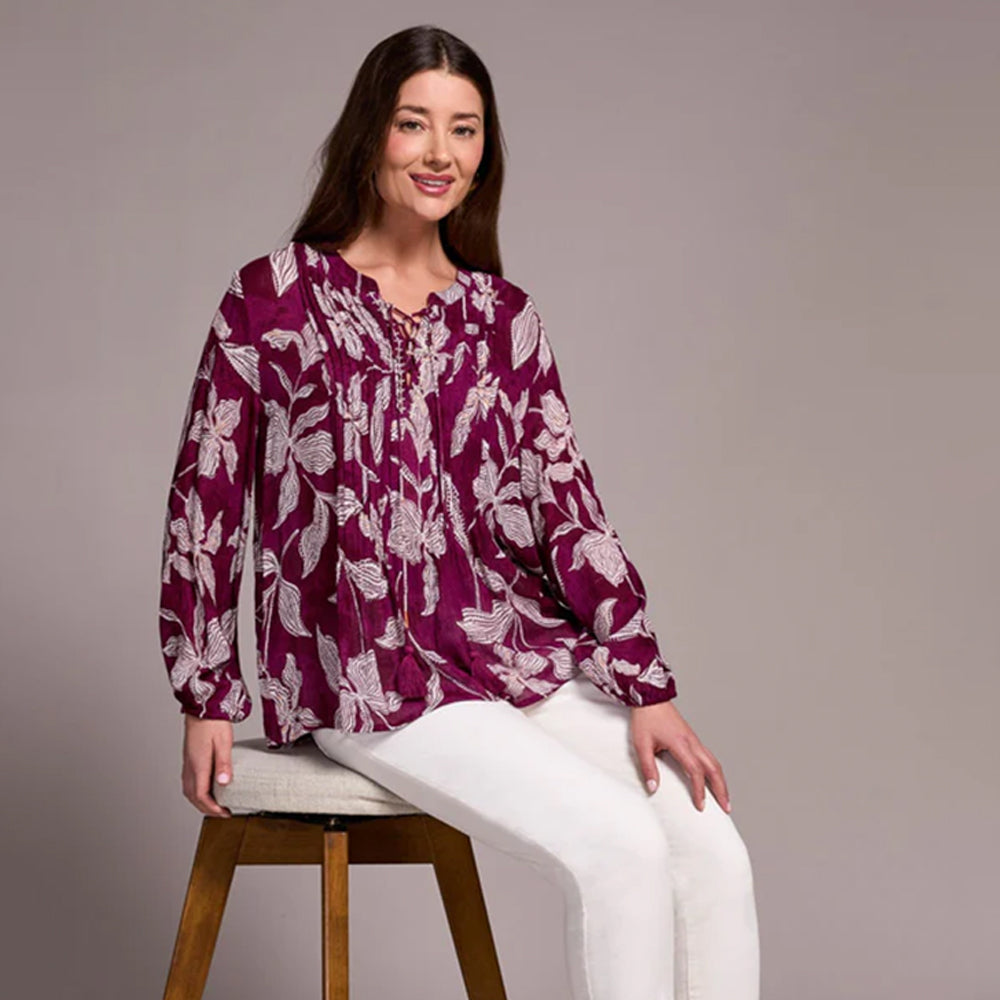 Woman wearing a purple floral blouse sitting on a wooden stool against a tan background