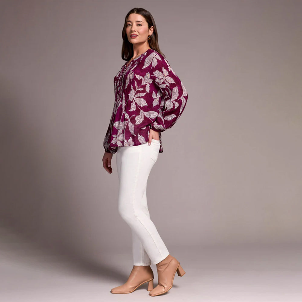 Woman wearing a purple floral blouse and white pants against a gray background