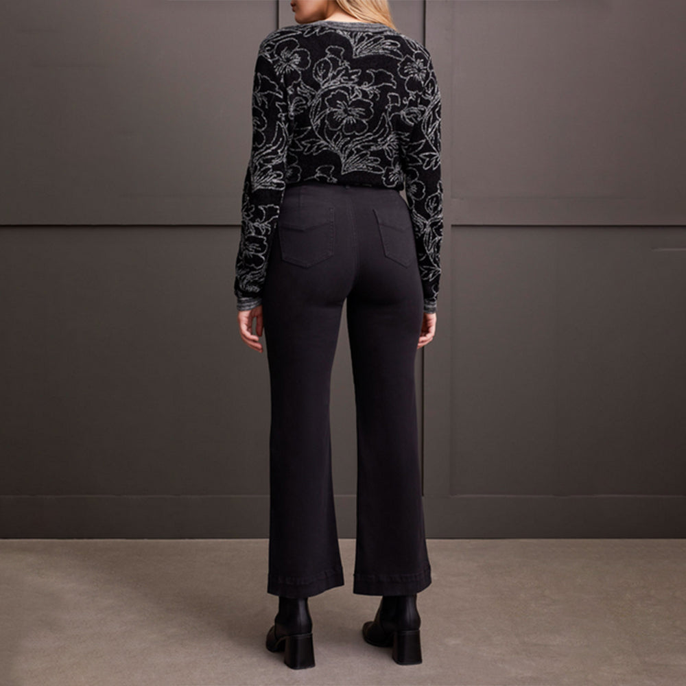 Person wearing a black patterned sweater and black pants against a gray wall.