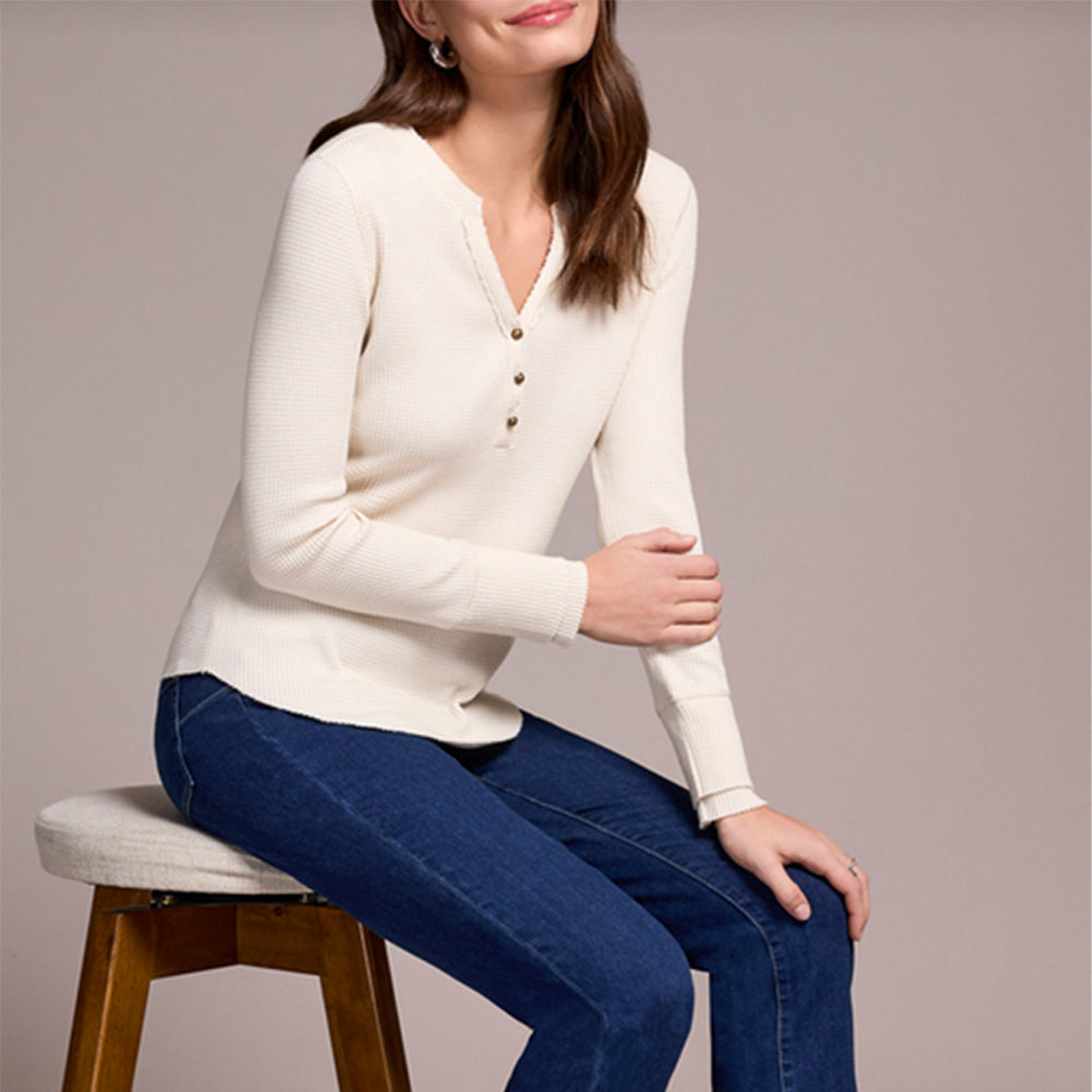 Woman wearing a white sweater and blue jeans sitting on a stool against a plain background