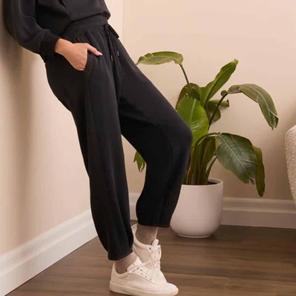 Person wearing black pants and white sneakers standing next to a potted plant indoors.