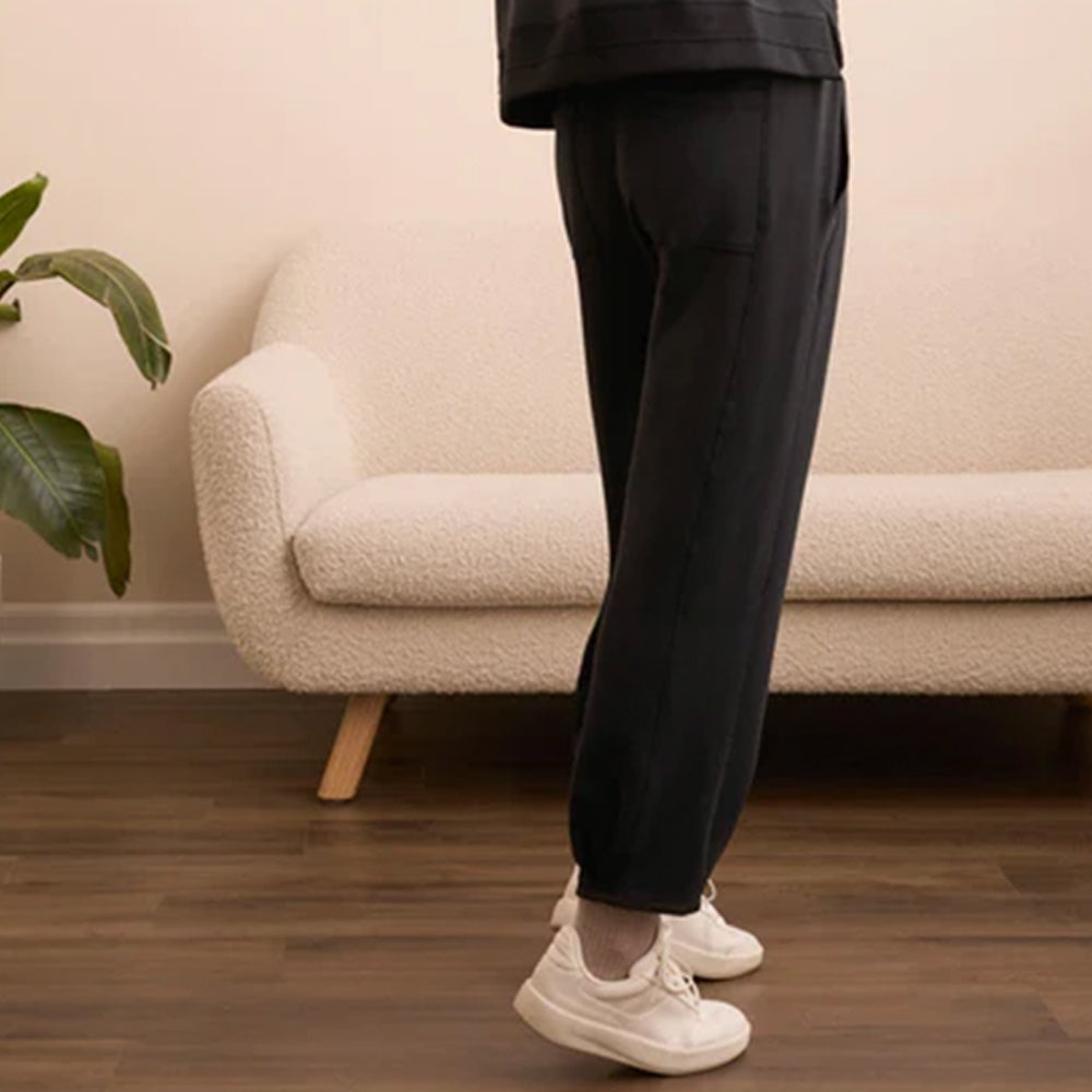 Person wearing black pants and white sneakers standing in a living room with a beige sofa and plant.