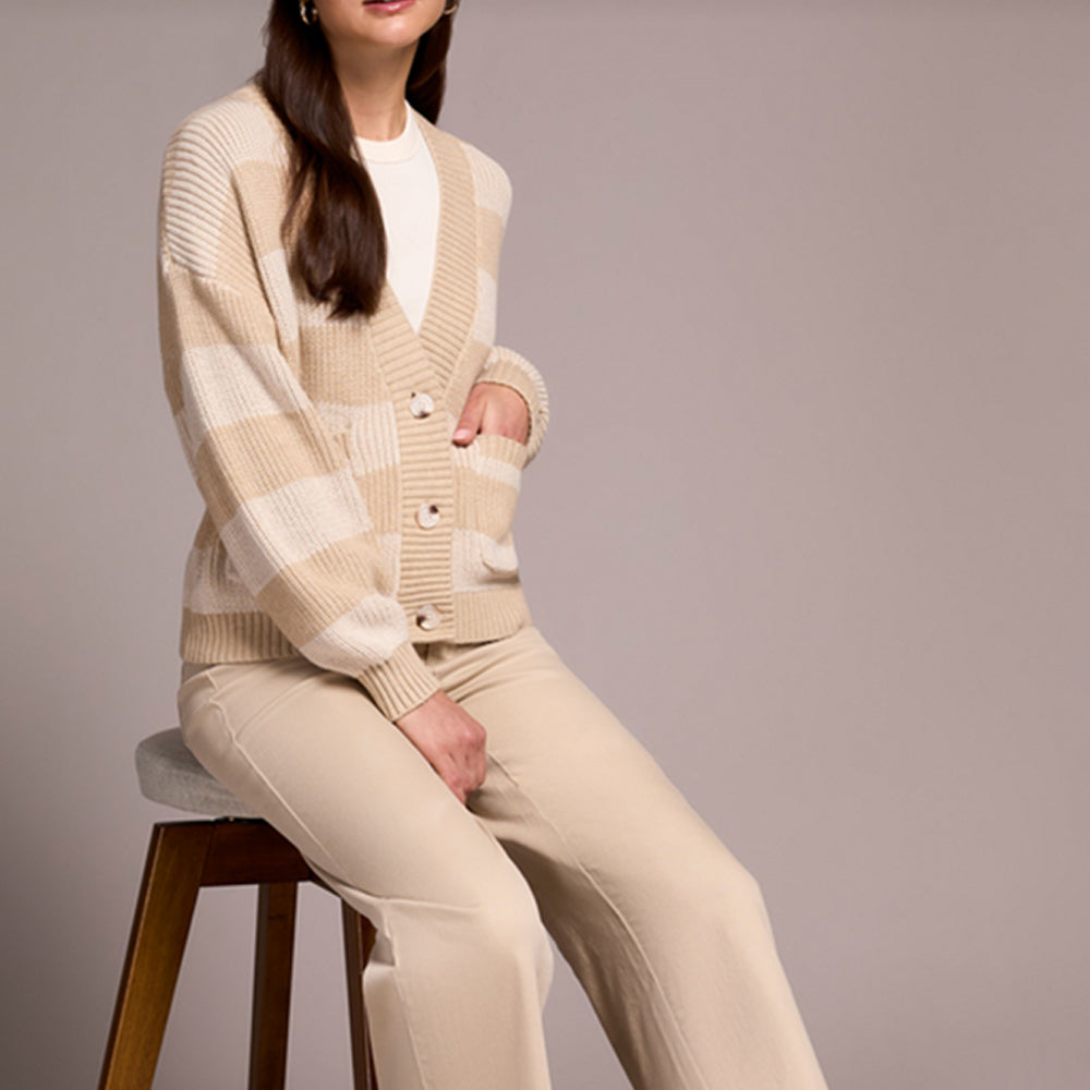 Woman wearing a beige cardigan and pants sitting on a stool against a plain background