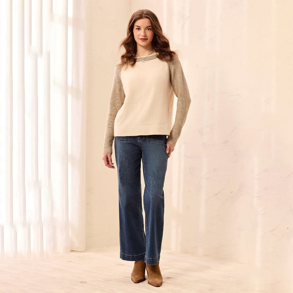 A woman modeling a beige knit sweater with textured taupe sleeves and a decorative collar, paired with dark blue wide-leg jeans and brown ankle boots, standing in a softly lit room with sheer curtains.