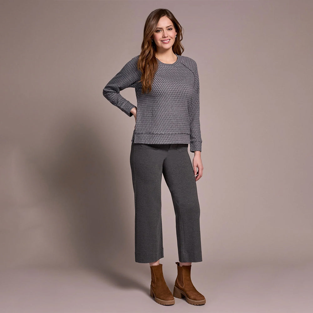 Woman wearing a gray patterned top and pants with brown boots against a beige background