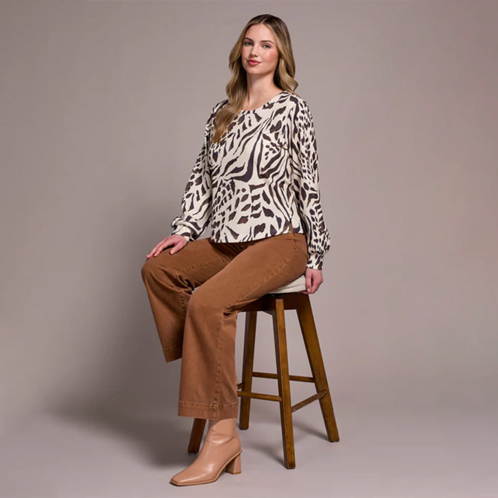 Woman sitting on a wooden stool wearing a patterned top and brown pants against a plain background