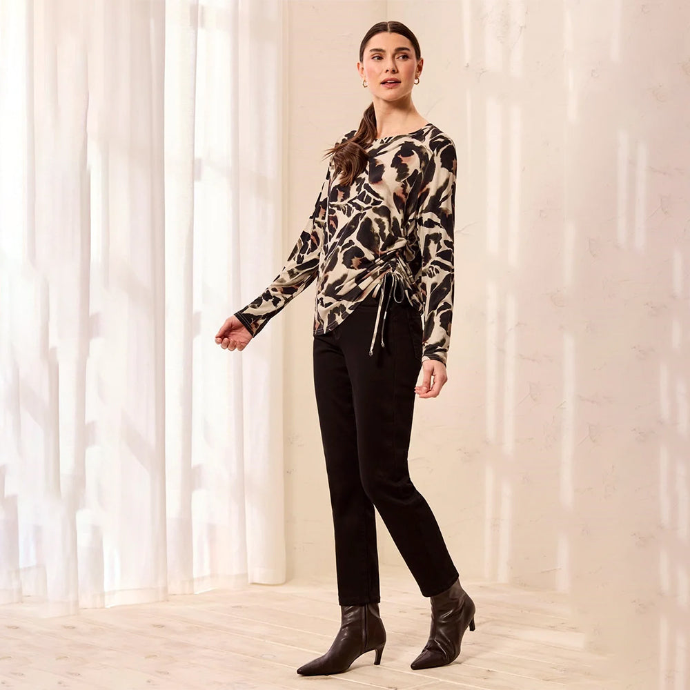A woman models a long-sleeve wrap-style top with an abstract black, brown, and beige pattern, paired with black pants and black ankle boots, standing in a brightly lit room with sheer curtains.