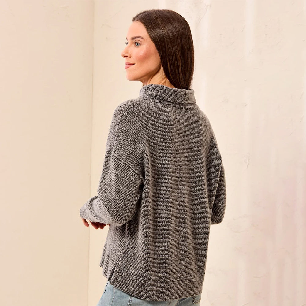 A woman wearing a textured gray knit mock neck sweater, shown from a three-quarter back view against a neutral cream-colored wall.
