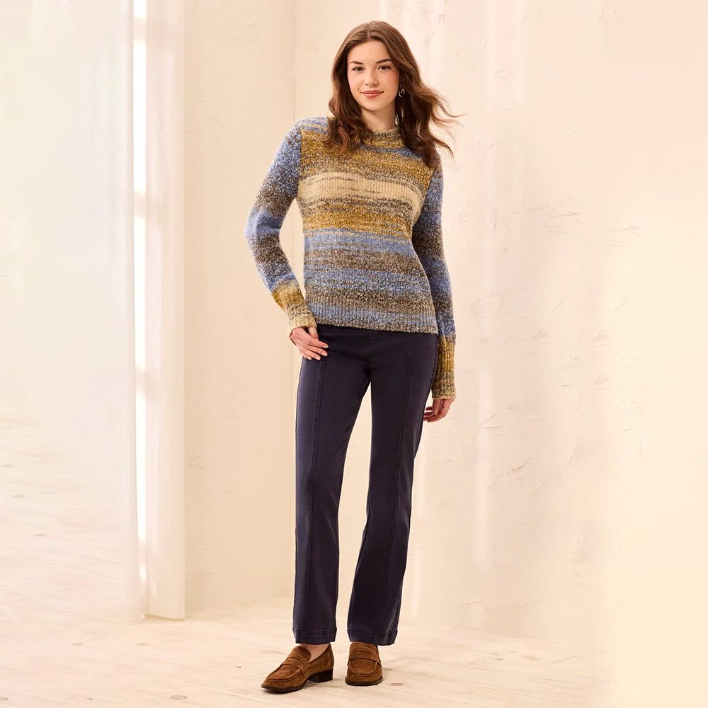 A woman stands in a bright indoor setting wearing a long-sleeve knit sweater with horizontal stripes in blue, yellow, and beige, paired with dark navy pants and brown suede loafers.