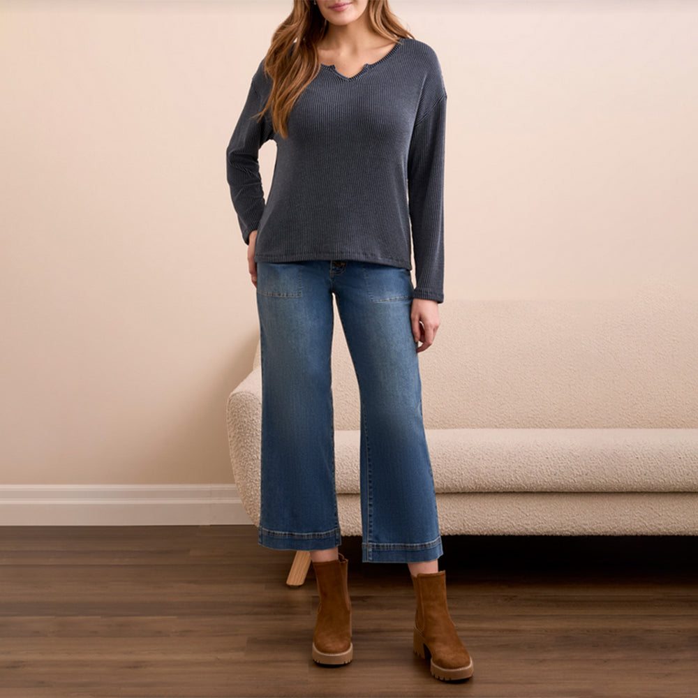 Woman wearing a dark gray long-sleeve top and blue jeans standing in a room with a beige wall and wooden floor.