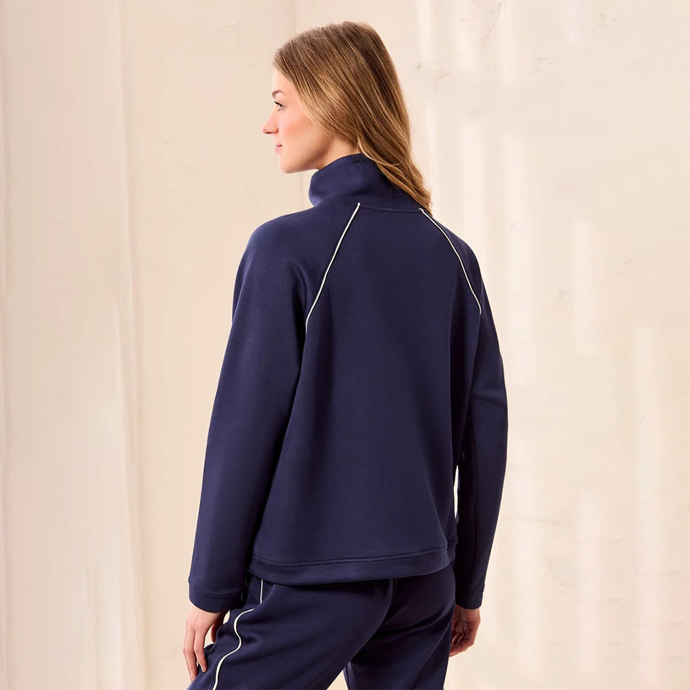 A woman shown from the back wearing a navy blue mock-neck athletic jacket with thin white piping details and matching navy pants, set against a neutral cream-colored wall.