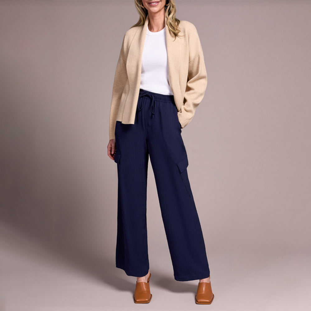 Woman wearing a beige cardigan, white shirt, navy pants, and brown shoes on a plain background