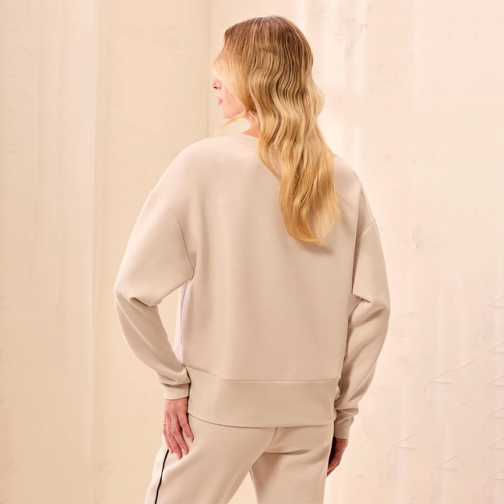 A woman with long blonde hair is shown from behind wearing a light beige crewneck sweatshirt and matching sweatpants against a soft, textured cream background.