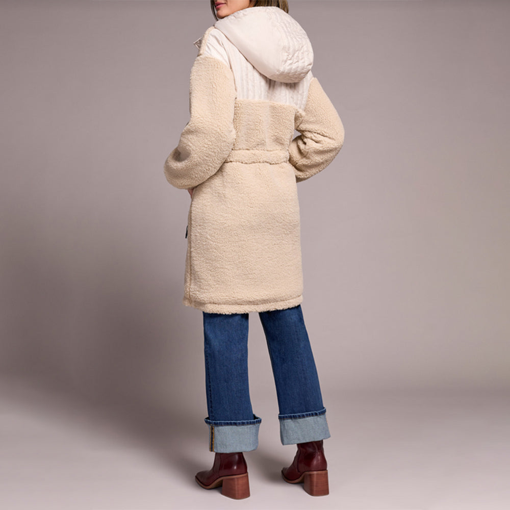 Person wearing a beige coat with a hood, blue jeans, and brown shoes on a plain background