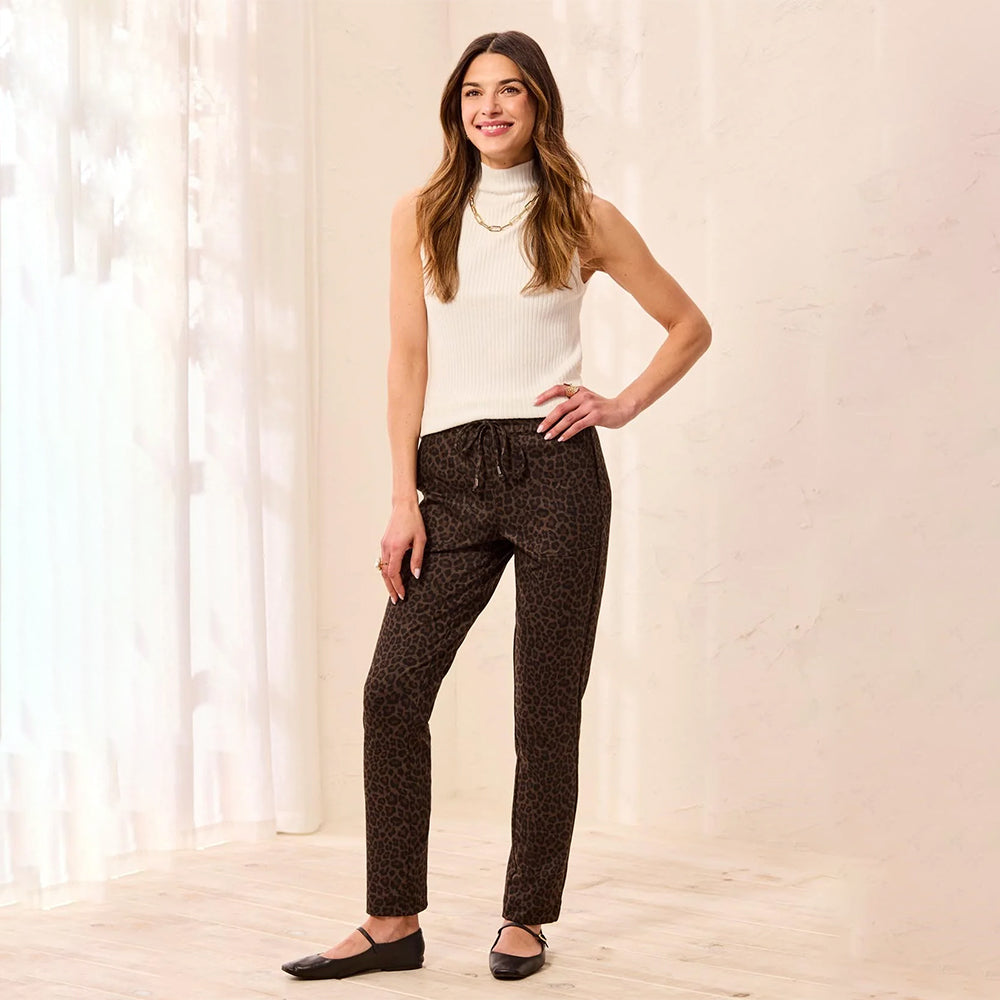 A woman modeling dark brown leopard-print pants with a drawstring waist, paired with a white ribbed sleeveless top and black flats, standing in a bright indoor setting with sheer curtains.