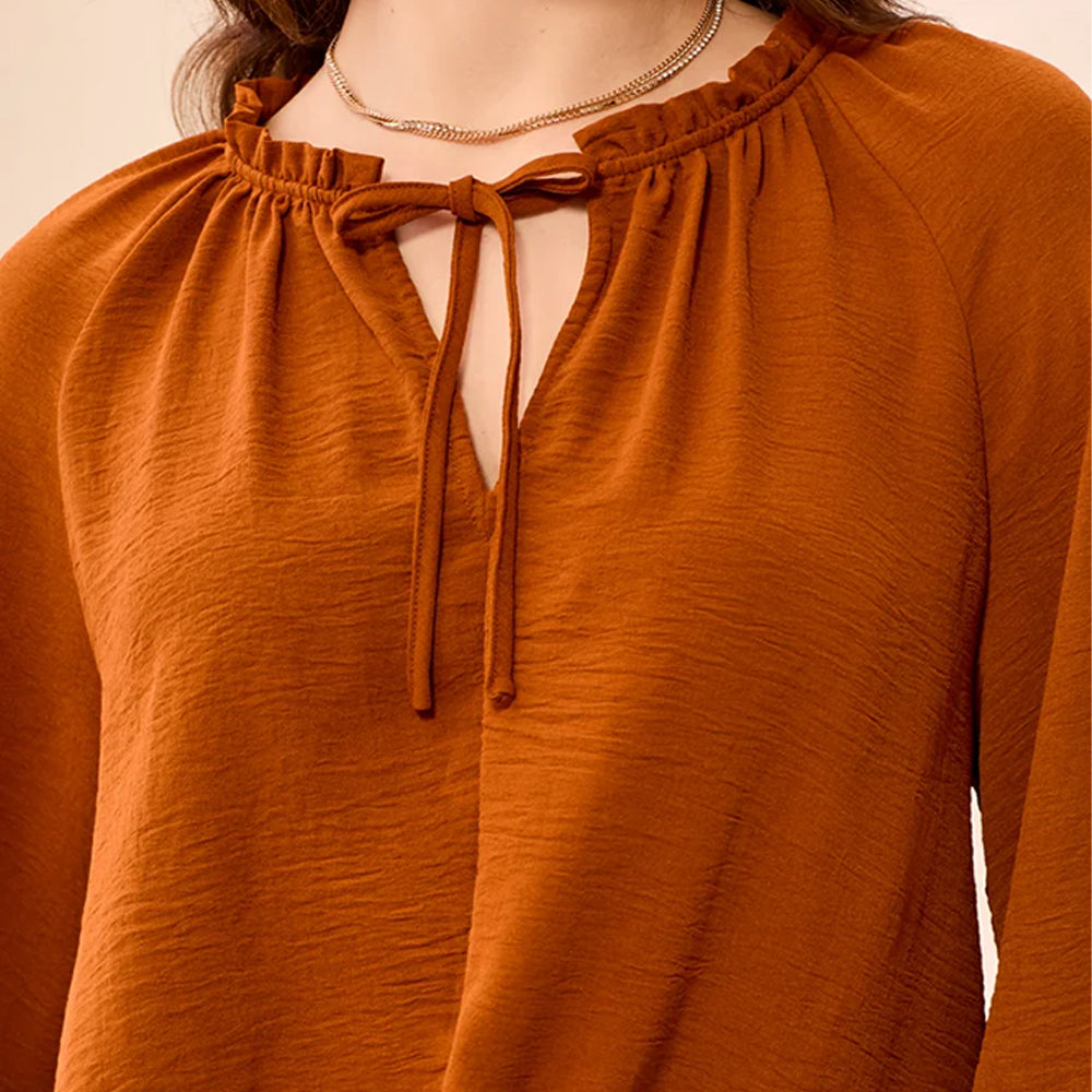 A close-up of a woman wearing a rust-orange long-sleeve blouse with a ruffled neckline and front tie detail, paired with layered gold necklaces.