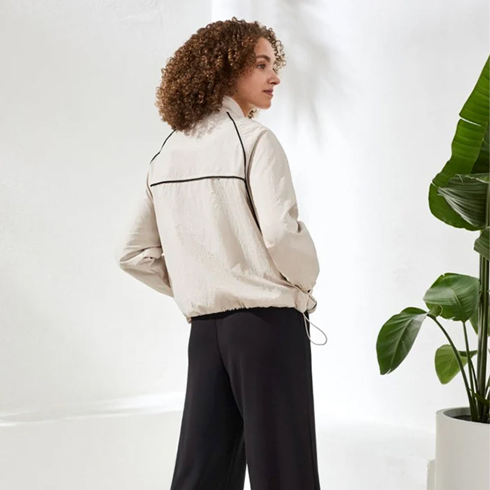 Woman wearing a beige jacket and black pants standing next to a potted plant indoors.