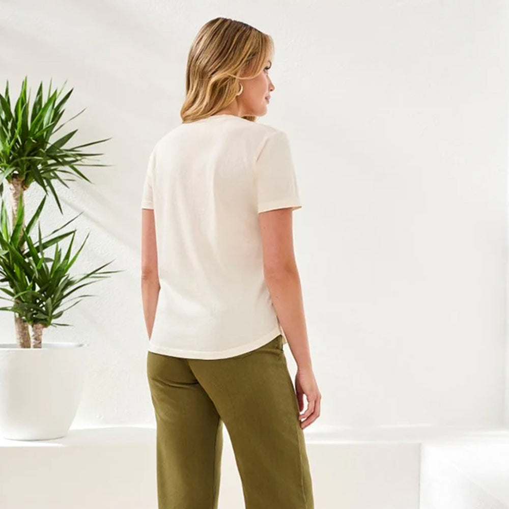 Woman wearing a white shirt and green pants standing in a minimalistic room with a plant.
