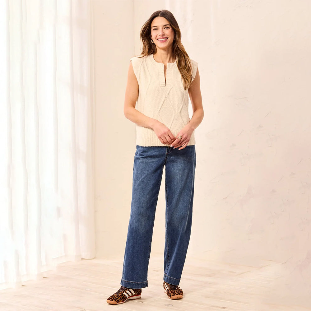 A woman modeling a sleeveless cream cable-knit vest with a split neckline, paired with wide-leg blue jeans and leopard-print sandals, standing in a bright, minimalist room with white curtains and a light-colored wall.