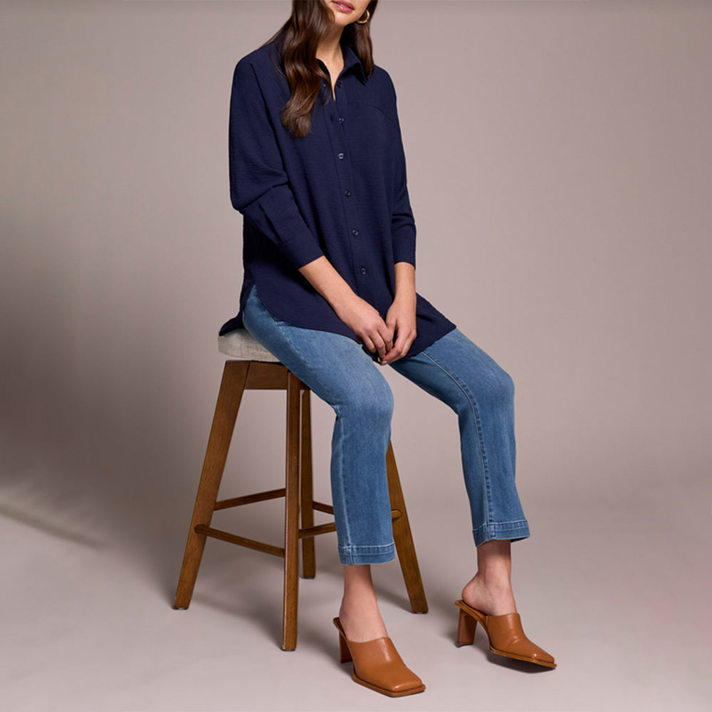 Person wearing a navy blue top and blue jeans on a beige background sitting on a stool.