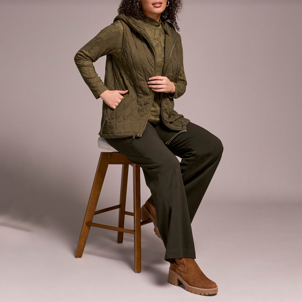 Woman wearing green zipper vest with brown pants and brown boots, sitting on a stool.