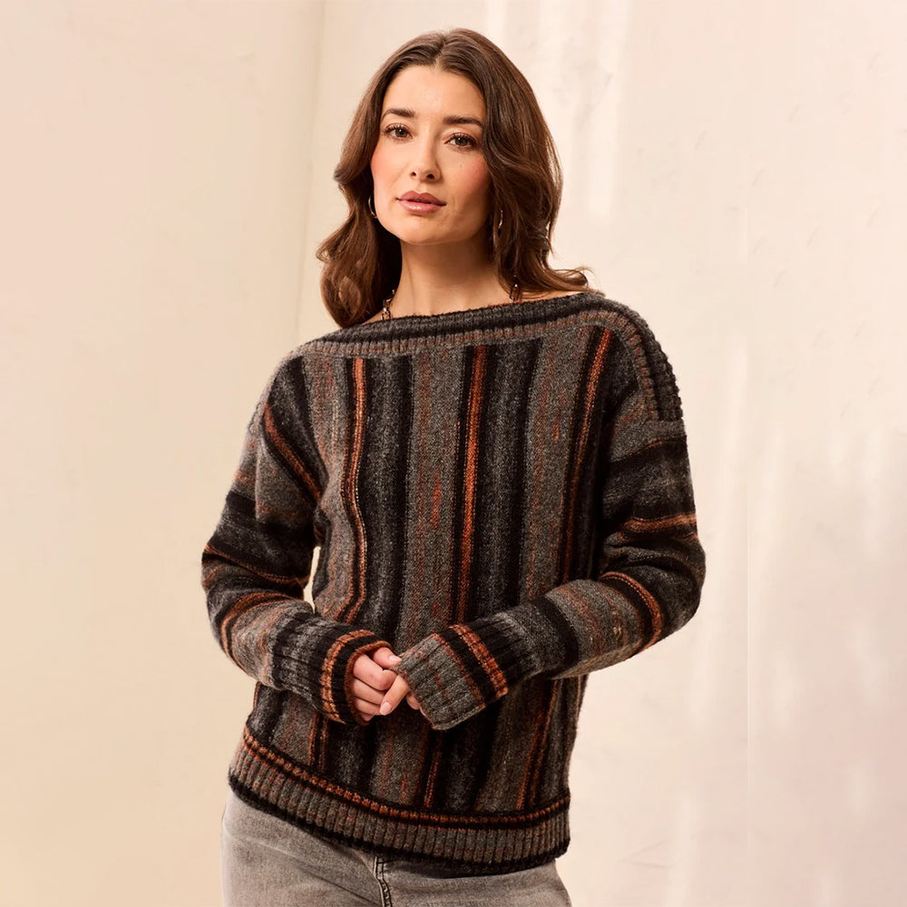 A woman wearing a dark gray and brown striped knit sweater with orange-brown vertical accents, paired with light gray jeans, standing against a neutral beige backdrop.