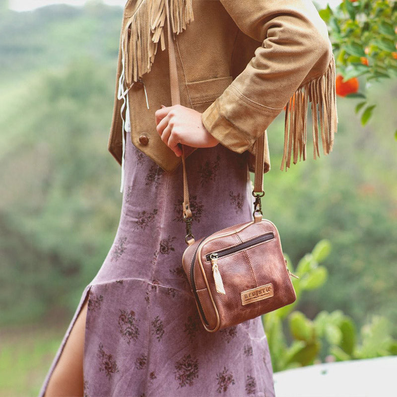 Bed Stu brown leather shoulder bag on the shoulder of a woman wearing a purple dress and a brown leather tassel jacket. 