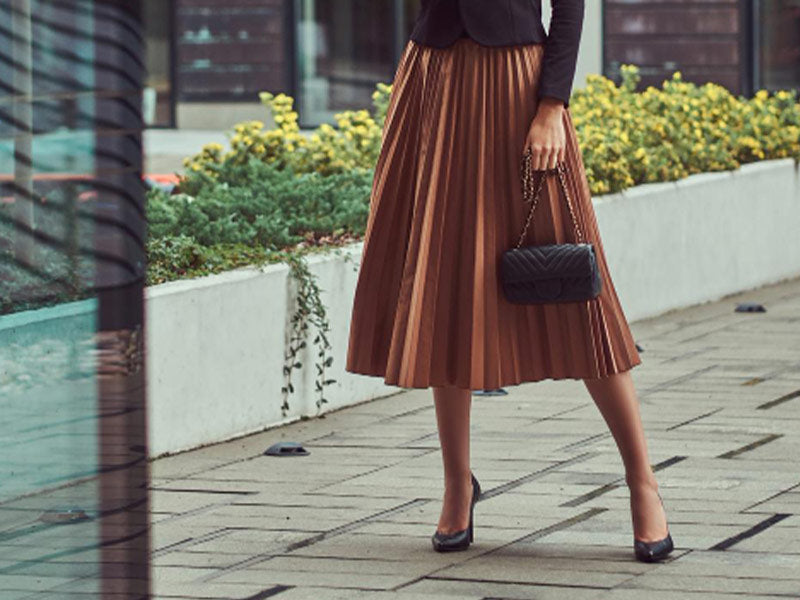 Woman wearing a black long sleeve shirt, a rust colored pleated knee high skirt, black heals, and holding a black hand bag.  