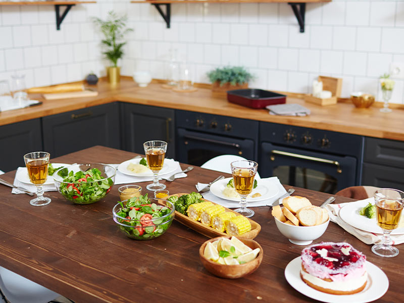 Picture of a kitchen island full of bread, salad, corn on the cobb, and wine glasses. 