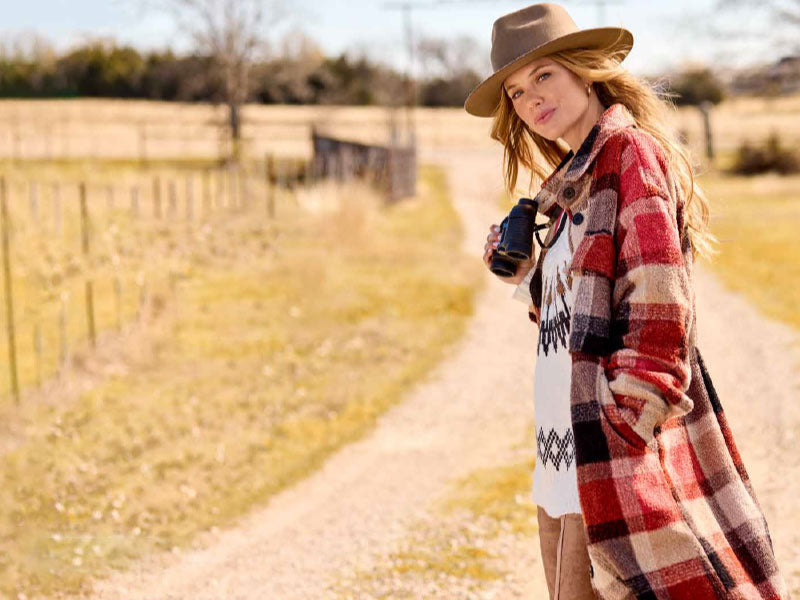 Girl standing on a dirt road wearing a tan hat and a long red flannel shacket and a tribal print top. 