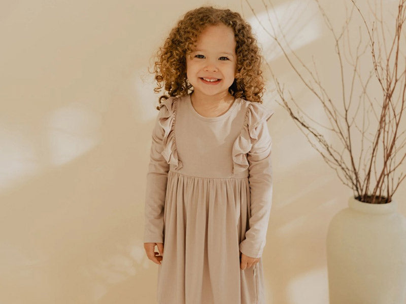Toddler girl with curly hair wearing a beige long sleeve dress with ruffles. 