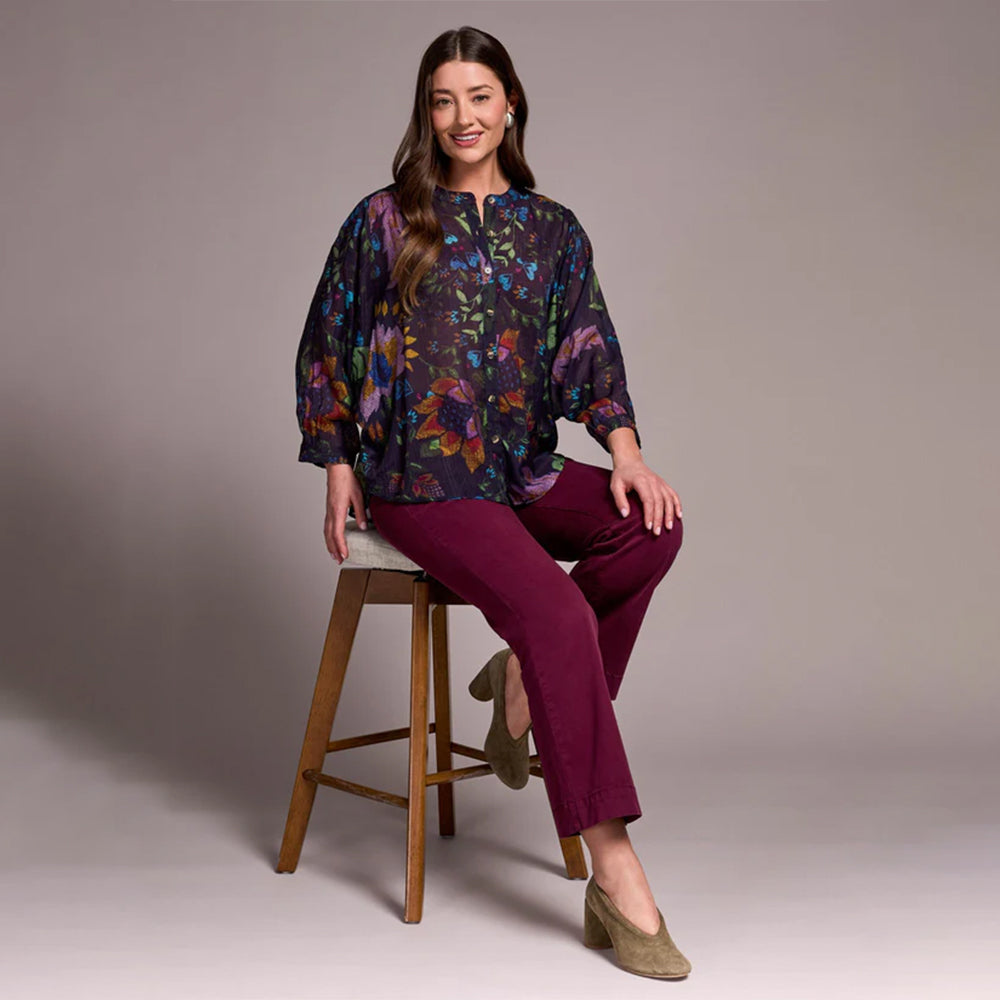 Woman sitting on a wooden stool wearing a colorful blouse and purple pants against a plain background