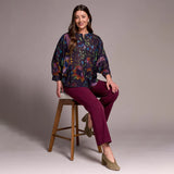 Woman sitting on a wooden stool wearing a colorful blouse and purple pants against a plain background